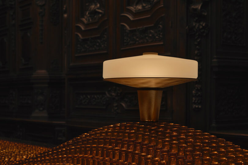 A modern, circular light fixture with a wide shade sits atop a bronze base, surrounded by numerous small cylindrical objects in a dark, ornate room.