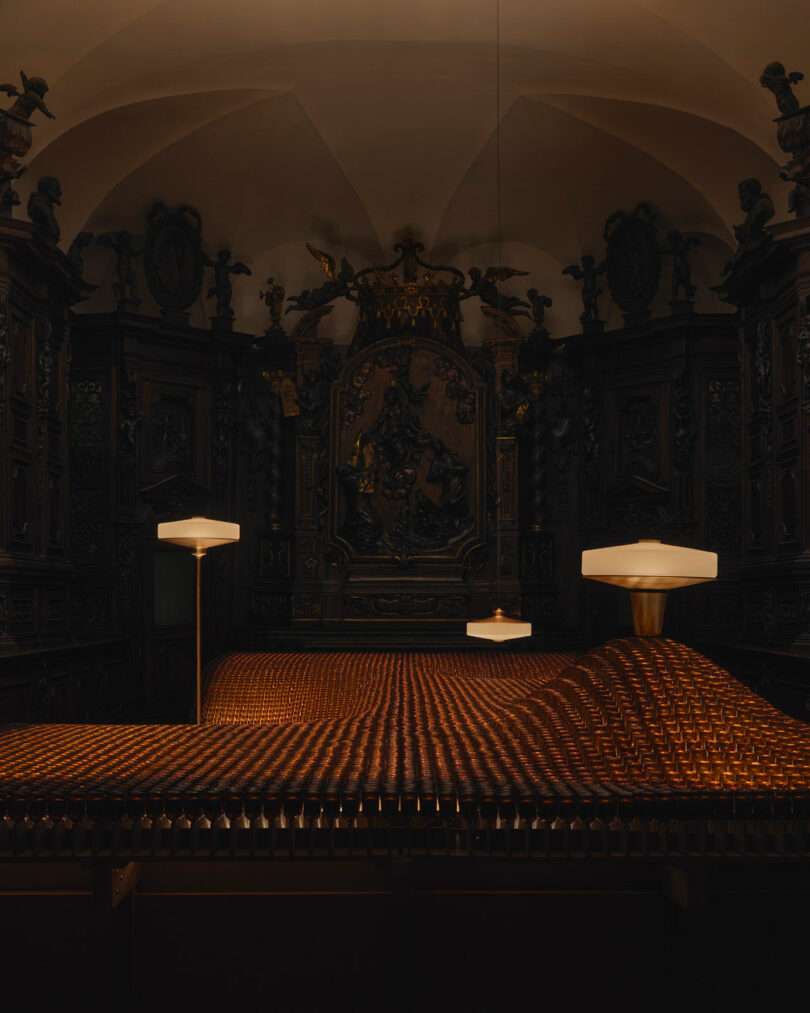 A dimly lit ornate room with elaborate wood carvings and a sculptural backdrop, featuring a modern installation of a wavy, grid-patterned surface and three contemporary pendant lights.