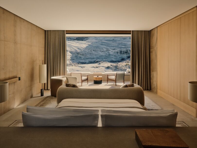 A modern, minimalist bedroom with neutral tones, a large bed, and seating area, featuring floor-to-ceiling windows overlooking a snowy mountain landscape.
