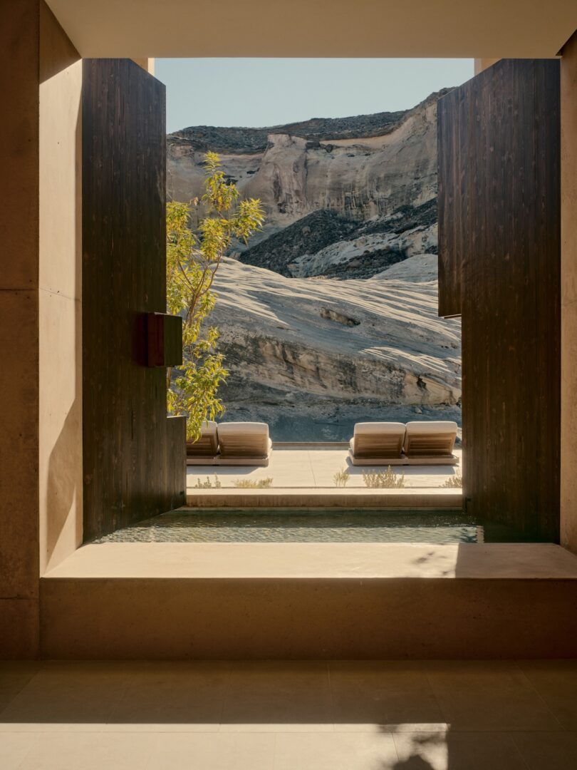 View through a modern stone and wood-framed opening to a small pool, lounge chairs, and rocky desert cliffs in the background.