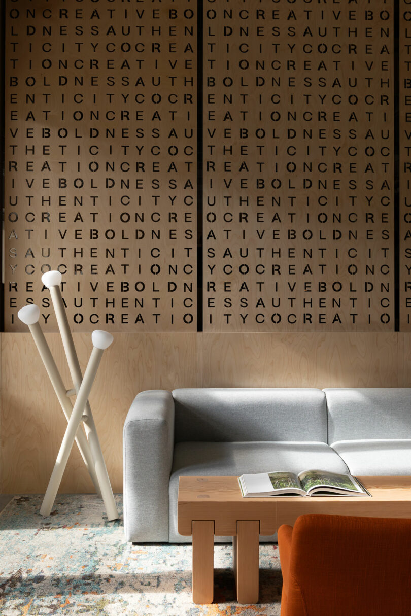 A modern living room with a gray sofa, wooden coffee table, open book, abstract floor lamp, and a wall featuring repeated bold words in a grid pattern.