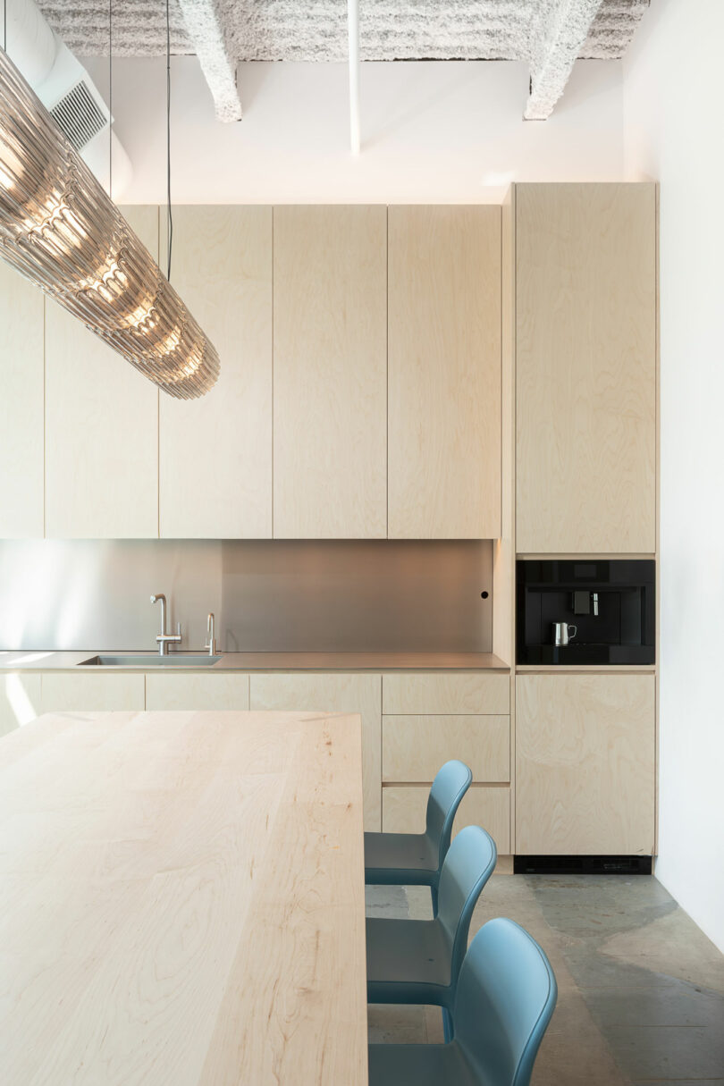 Minimalist kitchen with light wood cabinets, stainless steel backsplash, built-in coffee machine, and three blue chairs at a matching wood table under a modern pendant light.