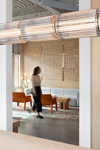 A person walks through a modern office lounge with orange chairs, a gray sofa, wooden accents, and a perforated wall panel with text.