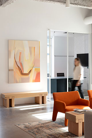 Modern office lounge with orange chairs, wooden bench, abstract art on the wall, and a person walking past a glass-walled meeting room.