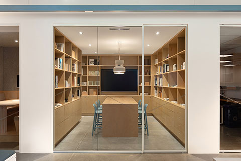 A modern meeting room with wooden shelves, a long wooden table, blue chairs, and a ceiling light, viewed through glass doors.