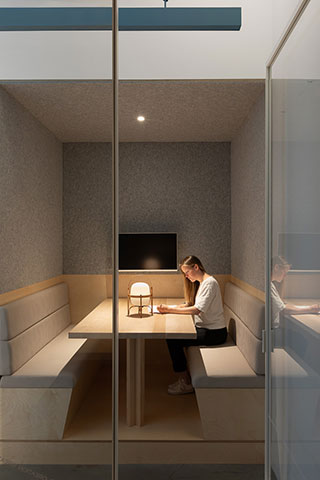 A woman sits alone at a table in a small, modern meeting room with padded benches, writing on paper under a desk lamp.