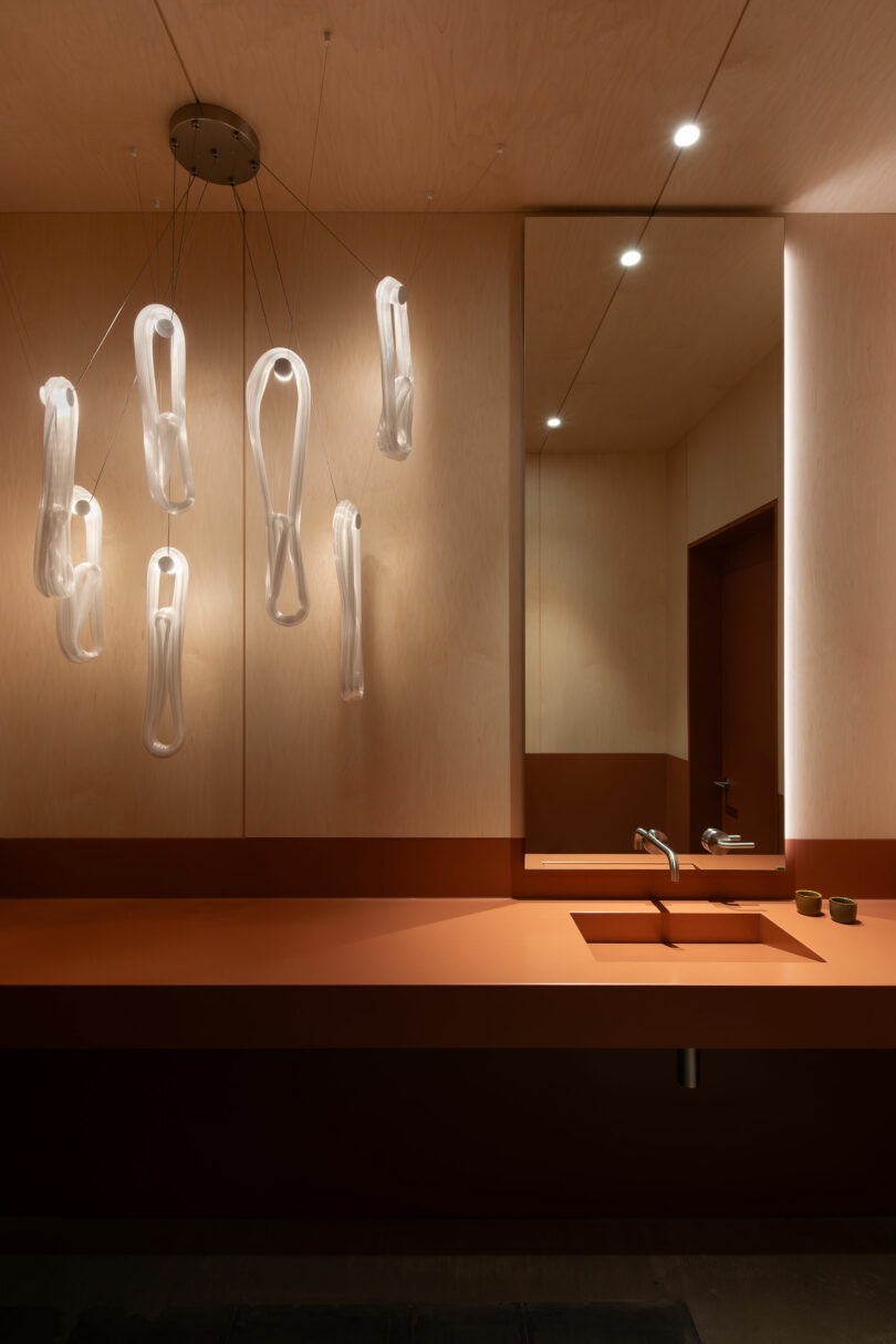 Modern bathroom with a long rust-colored countertop, integrated sink, large mirror, and sculptural glass pendant lights hanging from the ceiling.