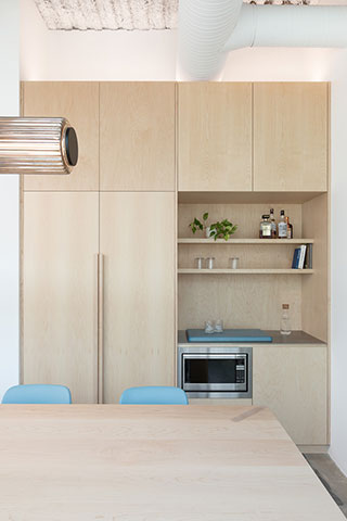 Minimalist kitchen with light wood cabinets, built-in shelves holding bottles and plants, a microwave, and a light wood dining table with blue chairs in the foreground.