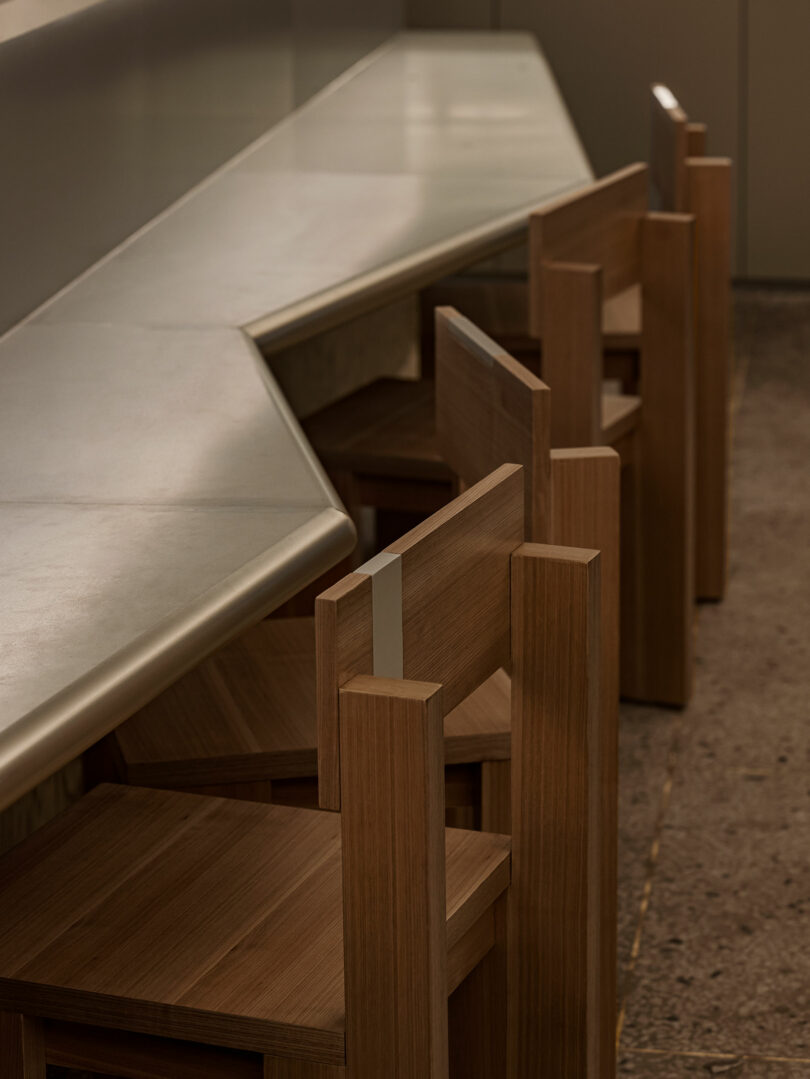 A row of wooden chairs lined up against a long, angular counter with a smooth, light surface in a minimalist interior setting.