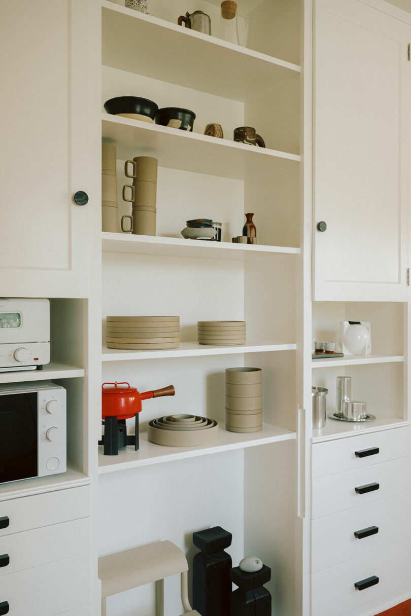 White kitchen shelves display neatly arranged dishes, bowls, containers, a red teapot, a microwave, and small kitchen appliances, all against a minimalist backdrop.