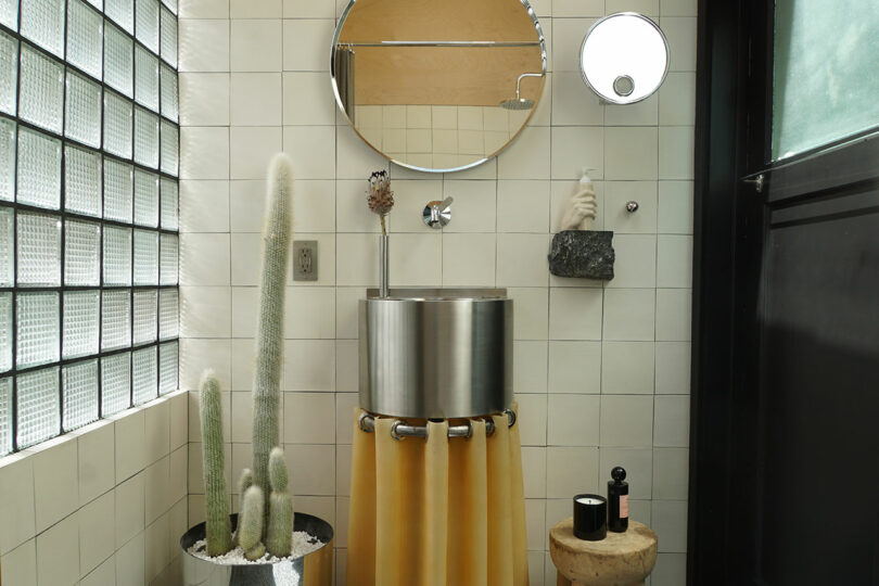 Modern bathroom with a round metal sink, circular mirrors, yellow curtain, cacti in a pot, stool with toiletries, and glass block wall.