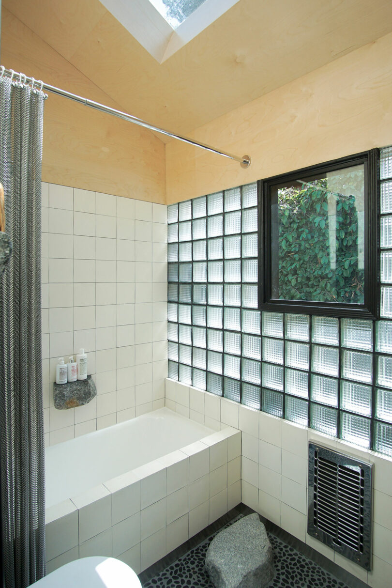 Modern bathroom with white tile bathtub, glass block wall, window, pebble stone floor, overhead skylight, metal curtain rod, and a small stone shelf holding toiletries.
