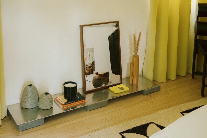 A low shelf with decorative vases, a candle, books, a framed mirror, a yellow notebook, and tall grass in a vase, set against a white wall with yellow curtains.