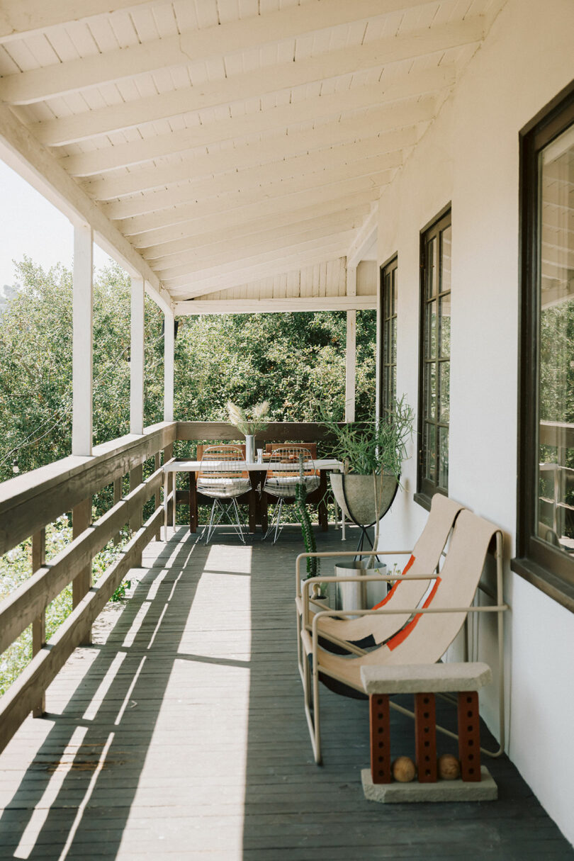 A wooden porch with a covered roof features two lounge chairs, a small table, and a dining area with four chairs and a table in the background, overlooking greenery.