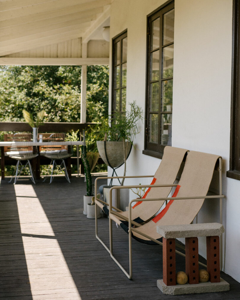 Two canvas lounge chairs with metal frames sit on a wooden porch next to potted plants, with a dining table and chairs visible in the background.