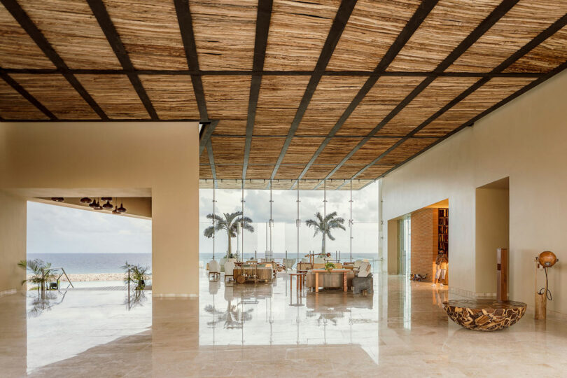 Spacious lobby with marble floors, wicker ceiling, modern furniture, glass wall, and ocean view framed by palm trees.
