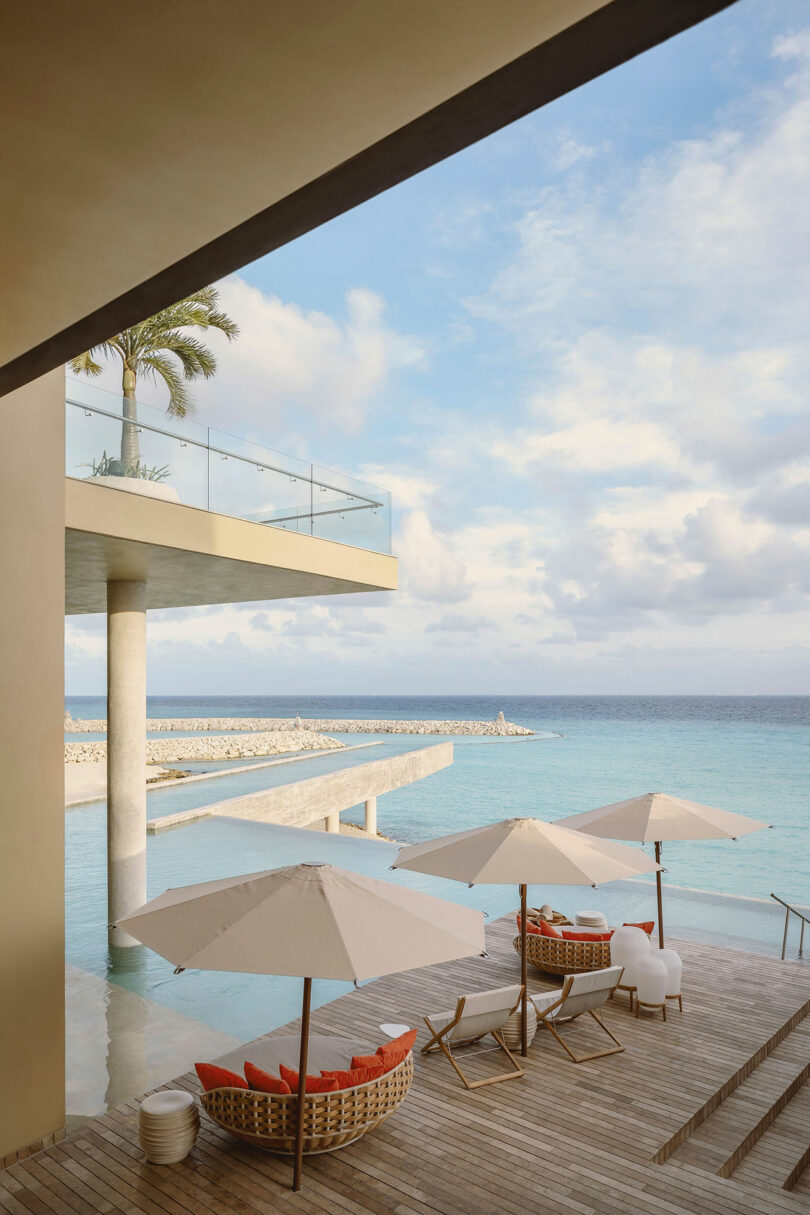 Modern resort terrace overlooking a blue ocean, with white umbrellas, lounge chairs, and a partially covered pool area under a sunny sky.