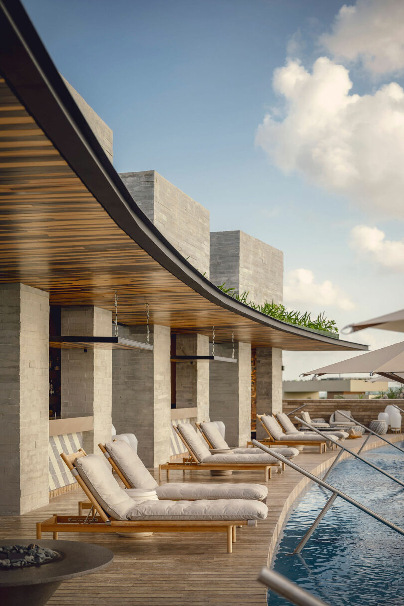 Modern outdoor pool area with cushioned lounge chairs on a wooden deck, shaded by large umbrellas, next to a contemporary stone building under a partly cloudy sky.