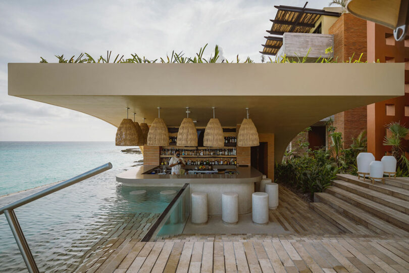 A modern, open-air bar with a thatched roof sits beside an infinity pool overlooking the ocean, surrounded by contemporary architecture and tropical plants.