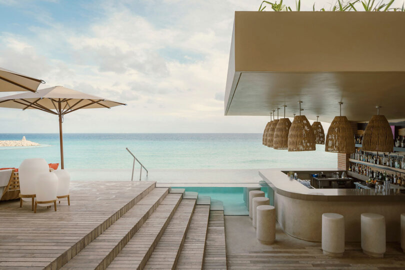 Modern beachfront bar with woven light fixtures, stool seating, wooden deck, and a small pool overlooking a calm, blue ocean under a partly cloudy sky.