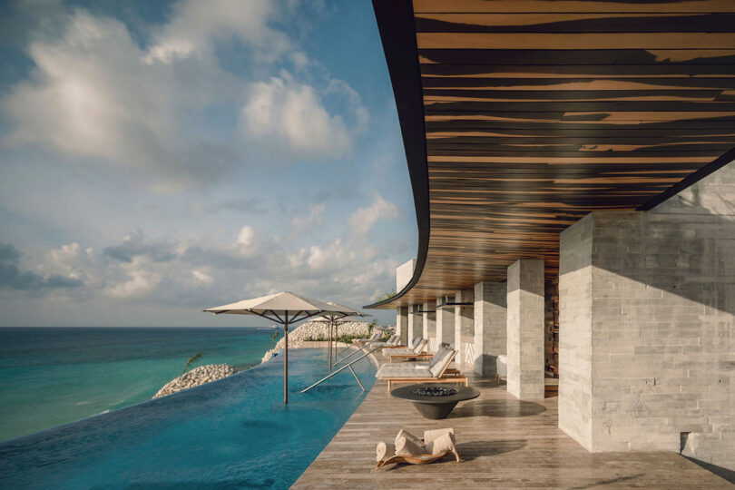 Infinity pool with lounge chairs and umbrellas beside a modern building overlooking the ocean under a partly cloudy sky.