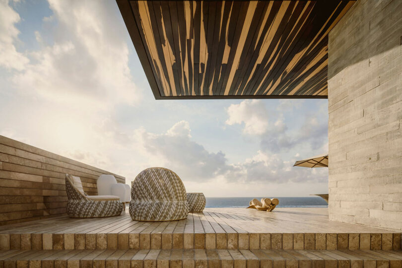 Modern outdoor patio with woven chairs on a wooden deck, overlooking the ocean under a partly cloudy sky, with architectural concrete elements.