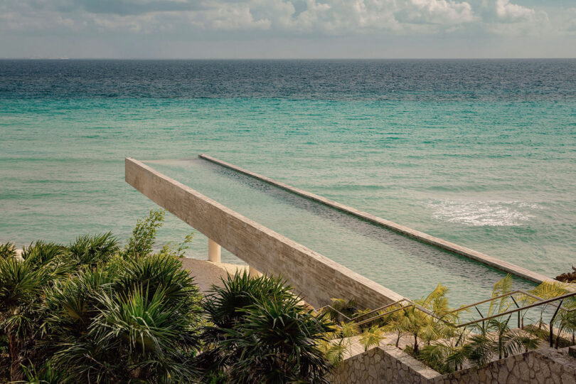 A long, narrow infinity pool extends toward the turquoise ocean, surrounded by tropical plants and stone steps under a partly cloudy sky.