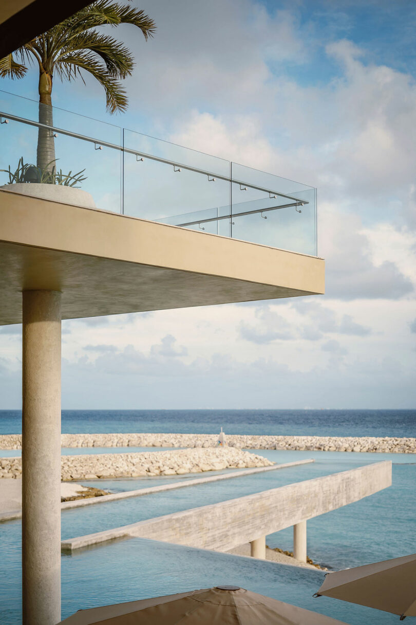 Modern building with glass balcony overlooking a pool and breakwater, with the ocean and sky in the background.