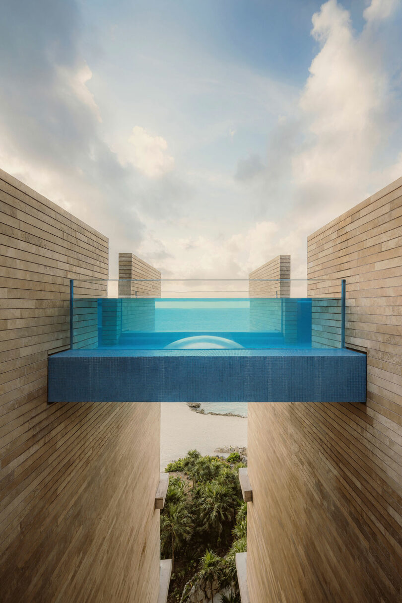 Rectangular glass-walled infinity pool suspended between two beige brick buildings, with a view of trees, beach, and sky in the background.