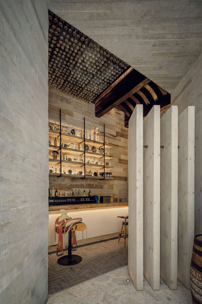 A modern bar with wooden paneling, open shelves stocked with bottles and decor, unique ceiling details, and two colorful saddle-shaped bar stools.