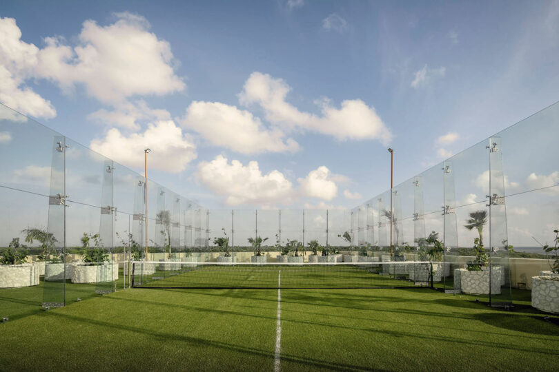 A rooftop sports court with glass walls, green artificial turf, white boundary lines, and potted plants, under a partly cloudy sky.