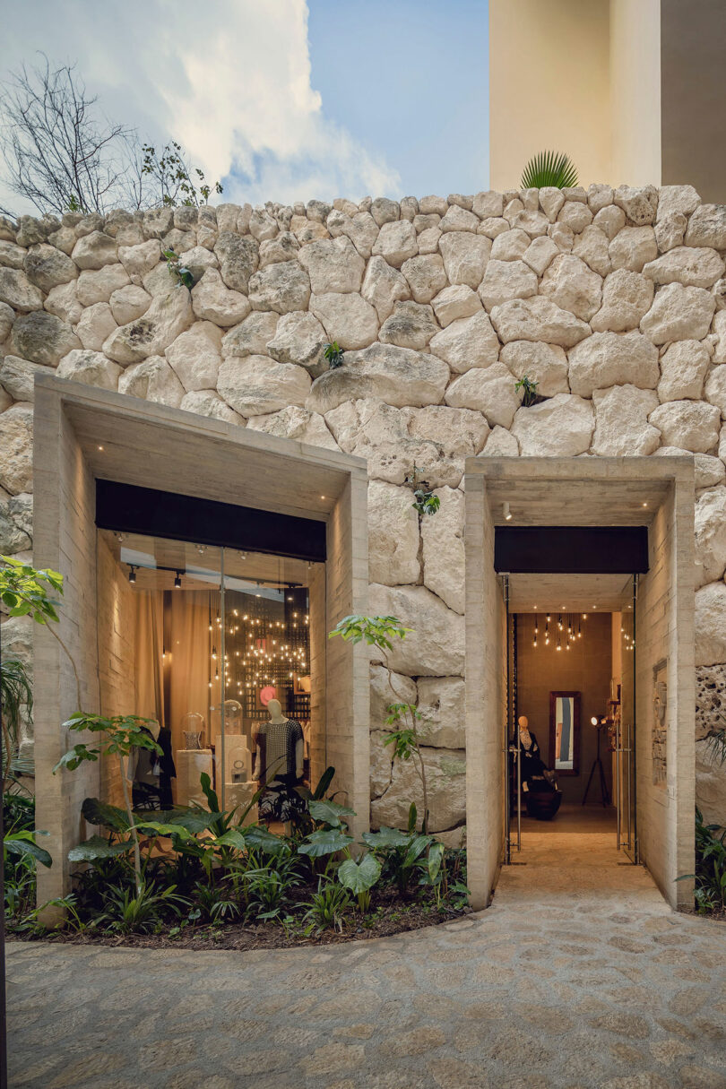 A modern entrance with two geometric doorways set in a stone wall, featuring large windows and hanging lights, surrounded by plants.
