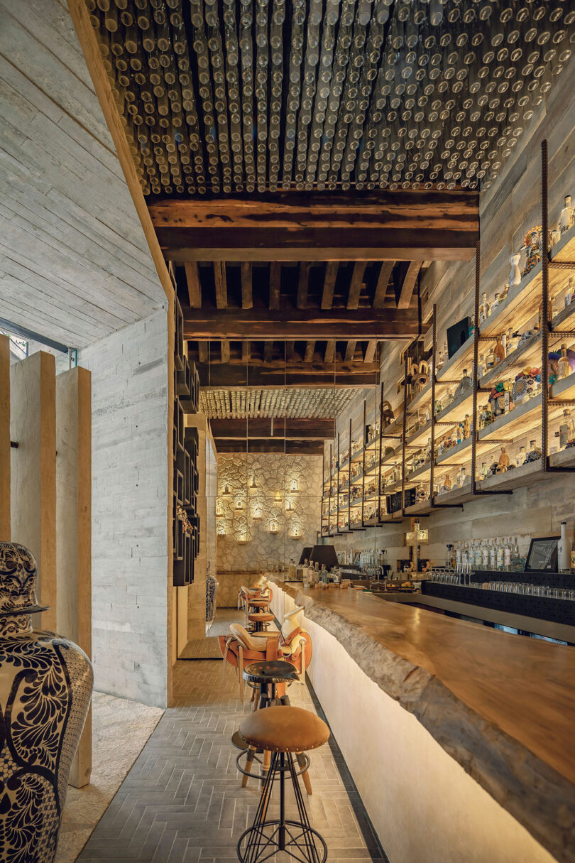 A modern bar with high wooden shelves holding bottles, a long wooden counter with stools, and a ceiling decorated with glass bottles.