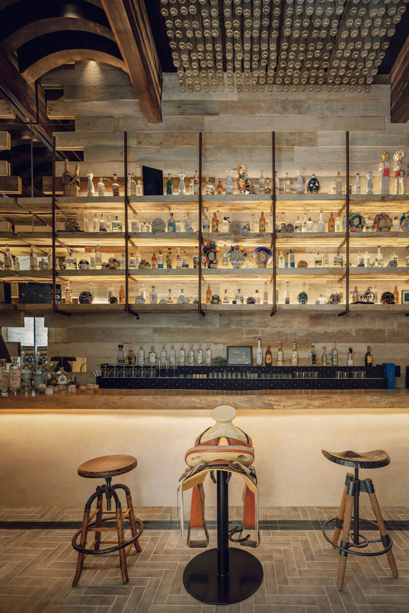 A bar with shelves displaying various bottles and decorative items above a counter, featuring three stools, including one designed with a saddle.