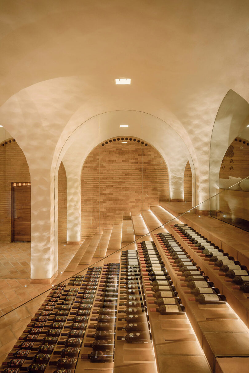 A modern wine cellar with arched ceilings, warm lighting, and rows of wine bottles displayed on angled shelves.