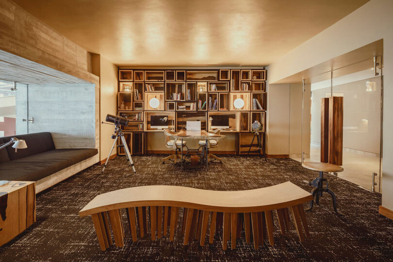 Modern office space with a wooden bookshelf, a central desk with computers, a telescope, a curved wooden bench, and glass-walled rooms on the side.