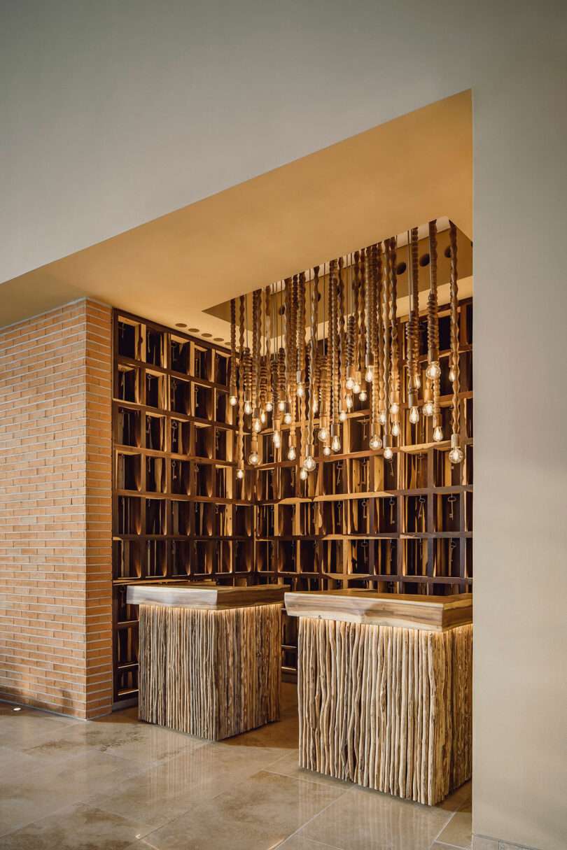 Modern reception area with two wood-textured desks, hanging exposed light bulbs, a brick wall on the left, and decorative shelving on the back wall.