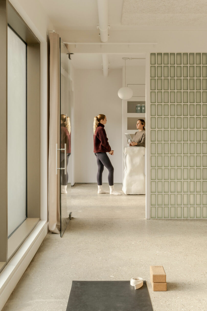 Two women stand and talk near a white reception desk in a bright, modern room with large windows, a yoga mat, and yoga blocks on the floor.