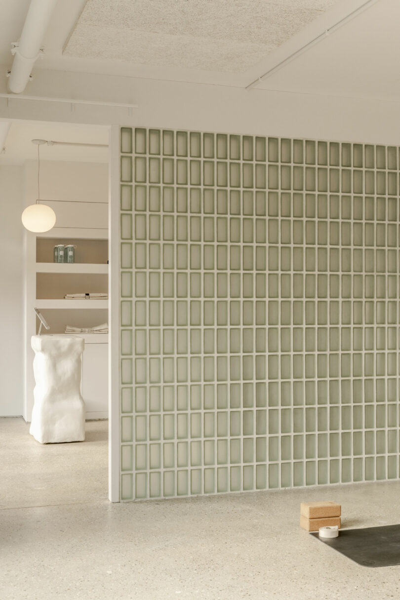 A room with a glass block partition wall, minimalist decor, white sculptural furniture, shelving with jars, and yoga props on a polished concrete floor.