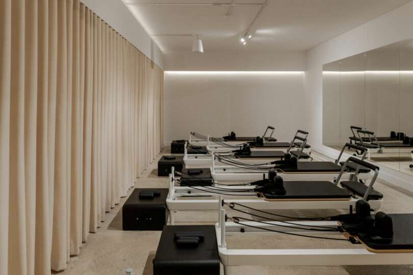 A modern Pilates studio with several reformer machines aligned in rows, beige curtains on one side, and a wall-length mirror on the other.