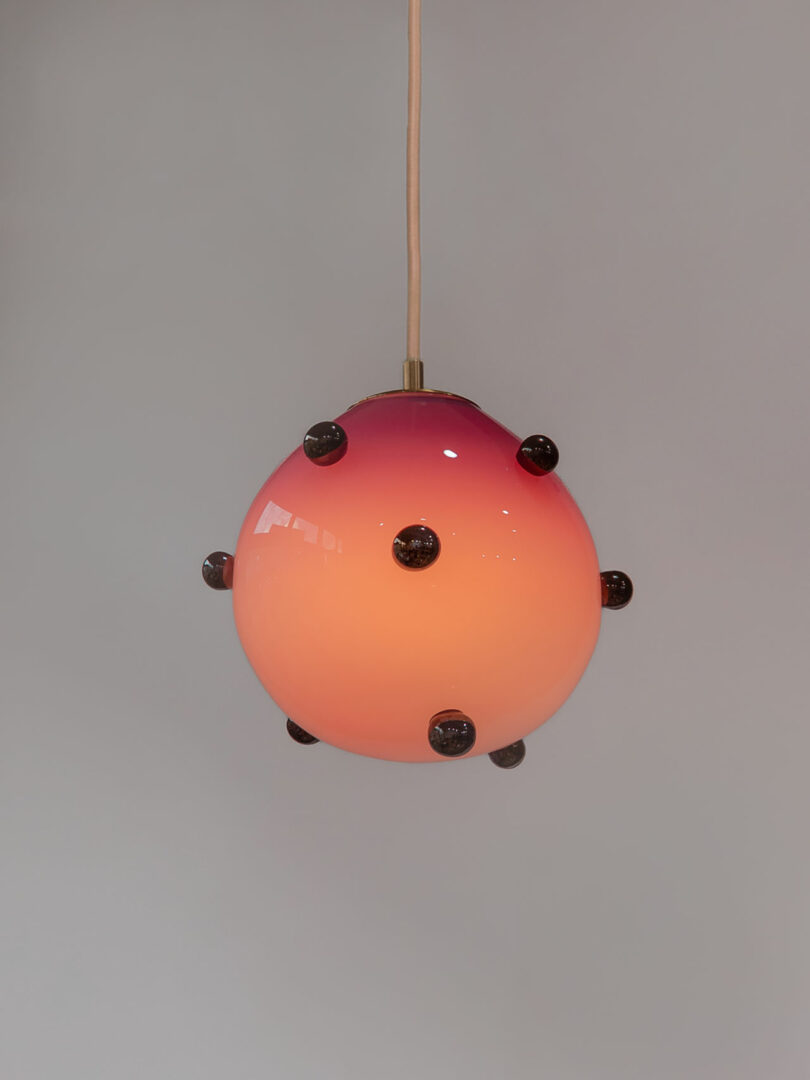 A spherical pendant light with a pink-to-orange gradient and multiple small dark circular accents hangs from a beige cord against a plain gray background.