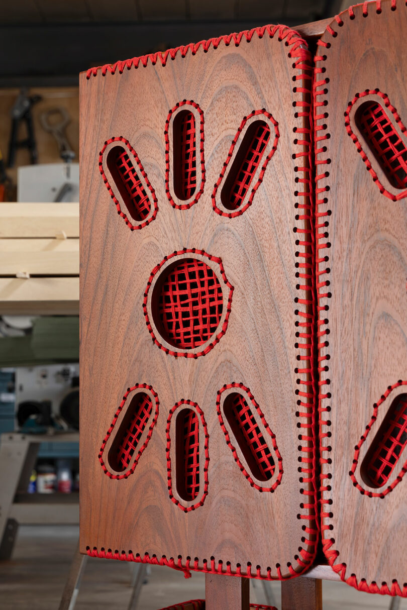A close-up of a wooden panel with red mesh and stitched edges, featuring cutouts in a sunburst pattern, in a workshop setting.