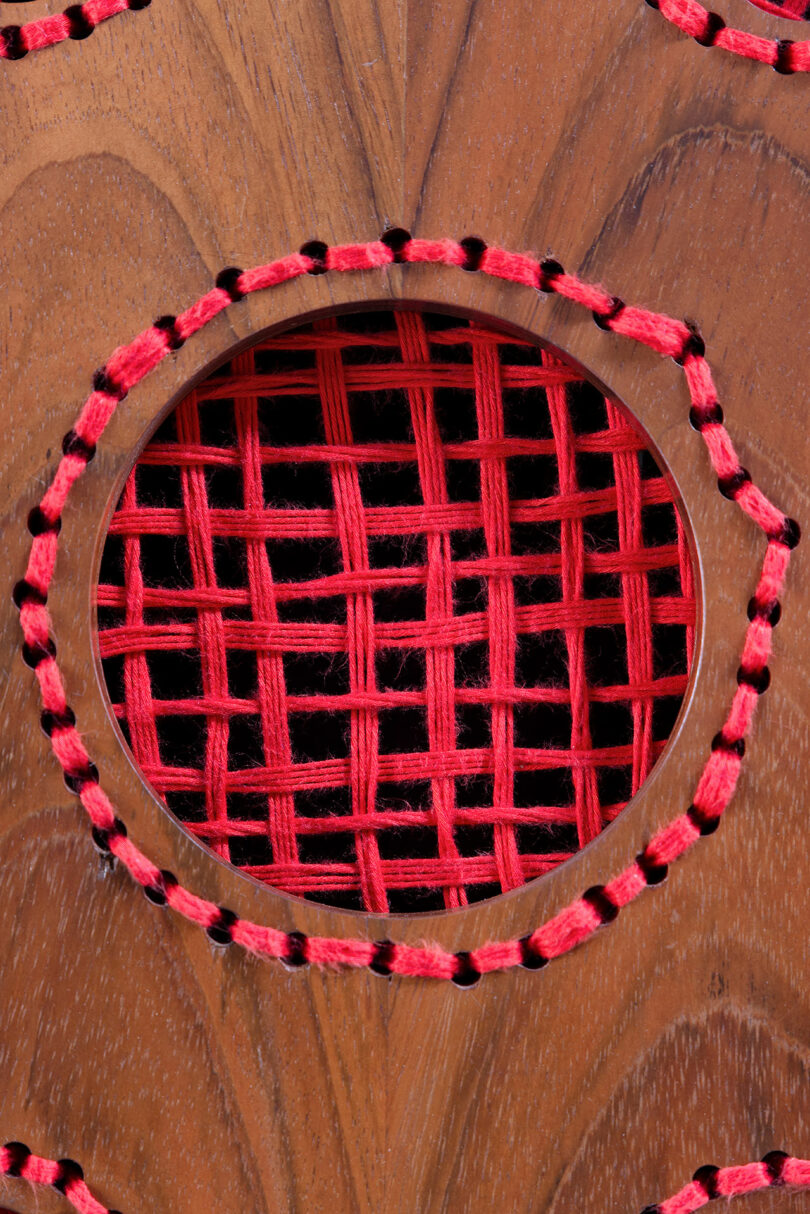 Close-up of a wooden surface with a circular opening bordered by black and red stitches, revealing a red woven pattern inside.
