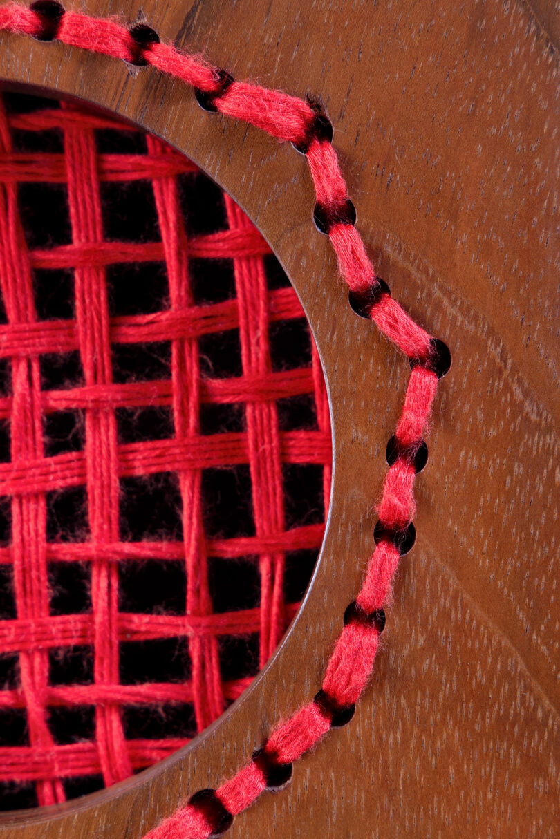 Close-up of red yarn woven in a grid pattern through a circular opening in a wooden surface, with additional yarn stitched along the edge of the opening.