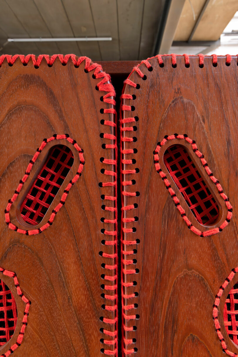 Close-up of two wooden panels with oval-shaped cutouts, joined together with visible red stitching; red lattice material fills the cutouts.