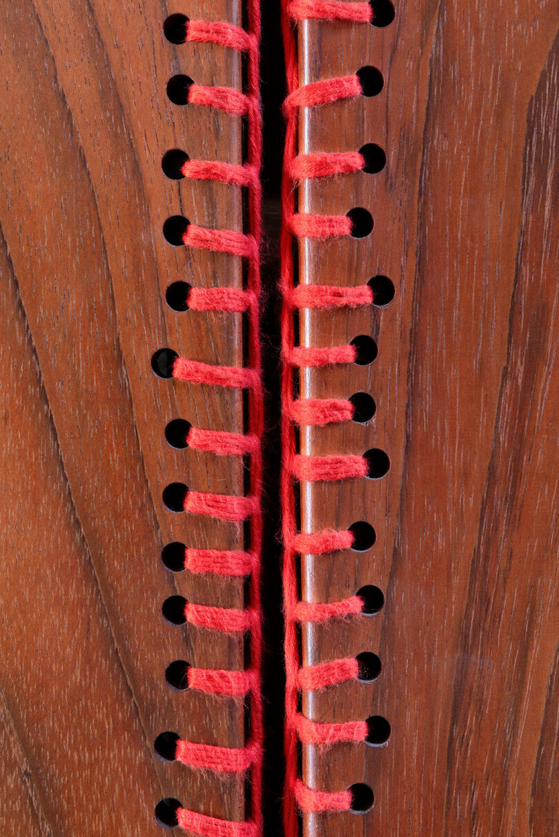 Close-up of two wooden panels joined together with red yarn threaded through evenly spaced holes along the edges.