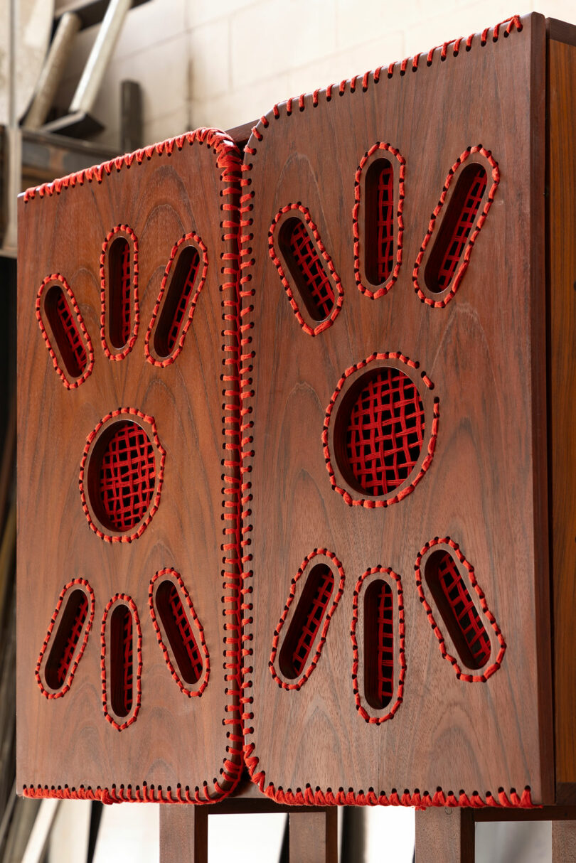 Wooden panels with red-stitched edges and cutouts in radial patterns, standing side by side in an indoor setting with tools and a ladder in the background.