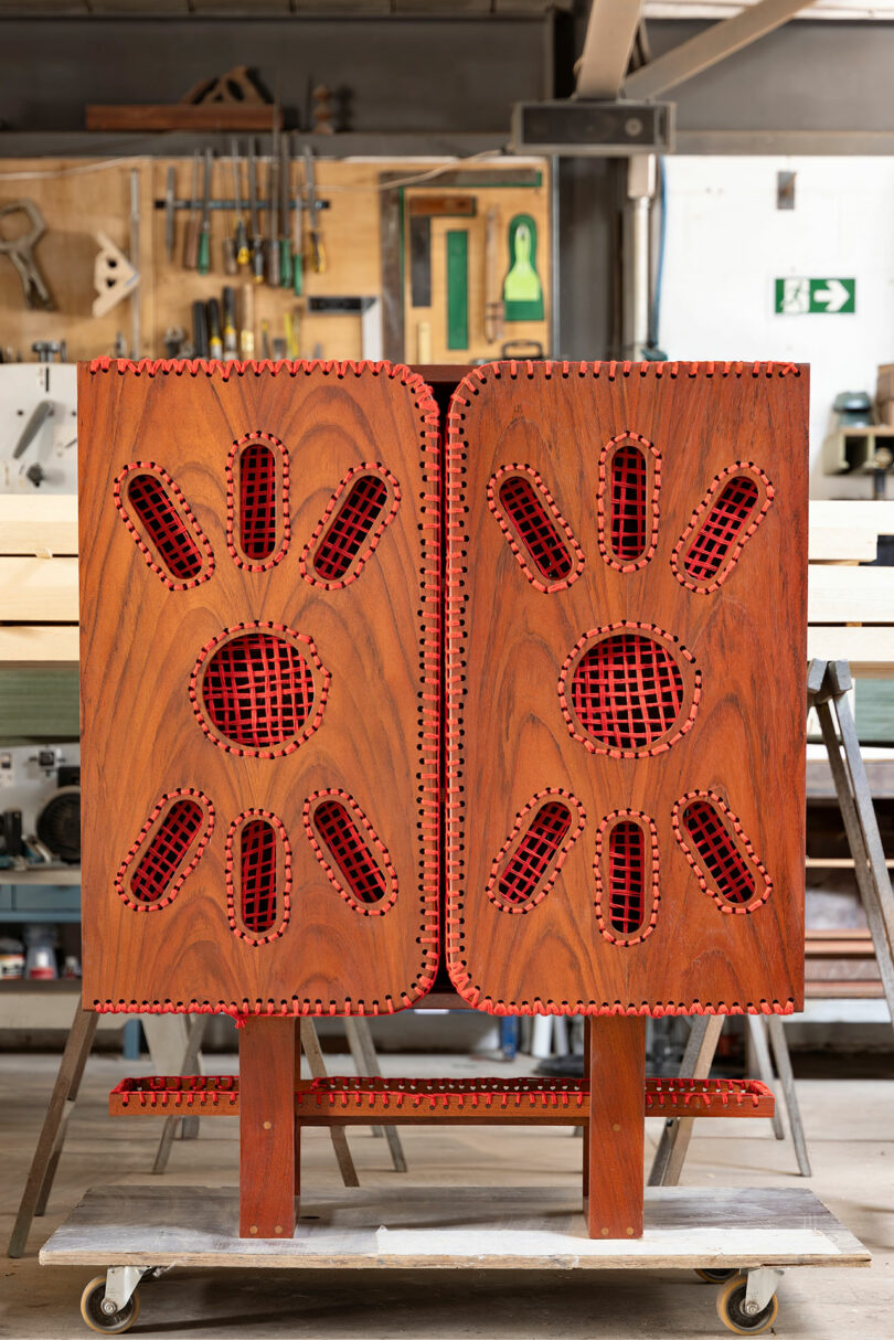 A wooden cabinet with decorative red mesh panels stands on a wheeled platform in a workshop with tools and wood planks in the background.