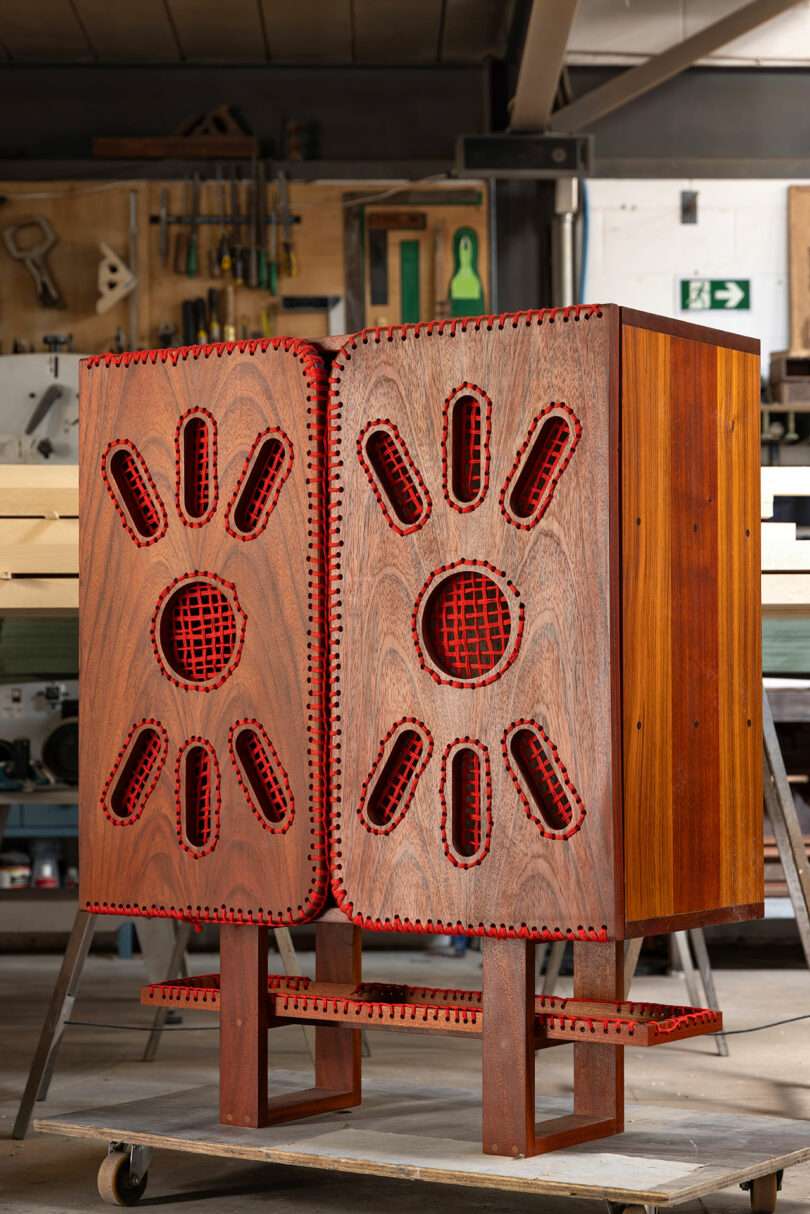 A wooden cabinet with intricate red-trimmed geometric cutouts stands in a workshop, surrounded by tools and woodworking equipment.