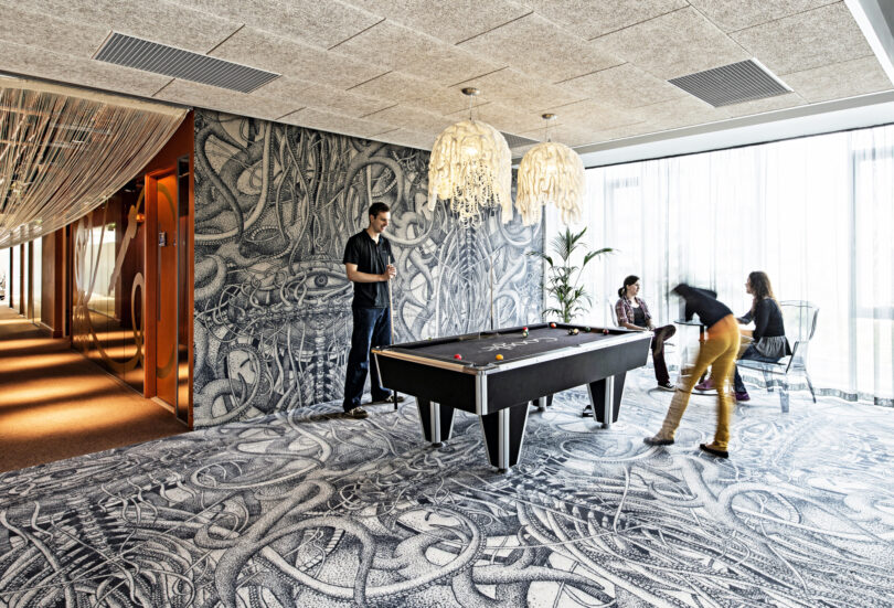 Four people enjoy a Friday 5 gathering in a modern room with artistic black-and-white wall and floor designs, playing and watching a game of pool on a sleek black pool table.